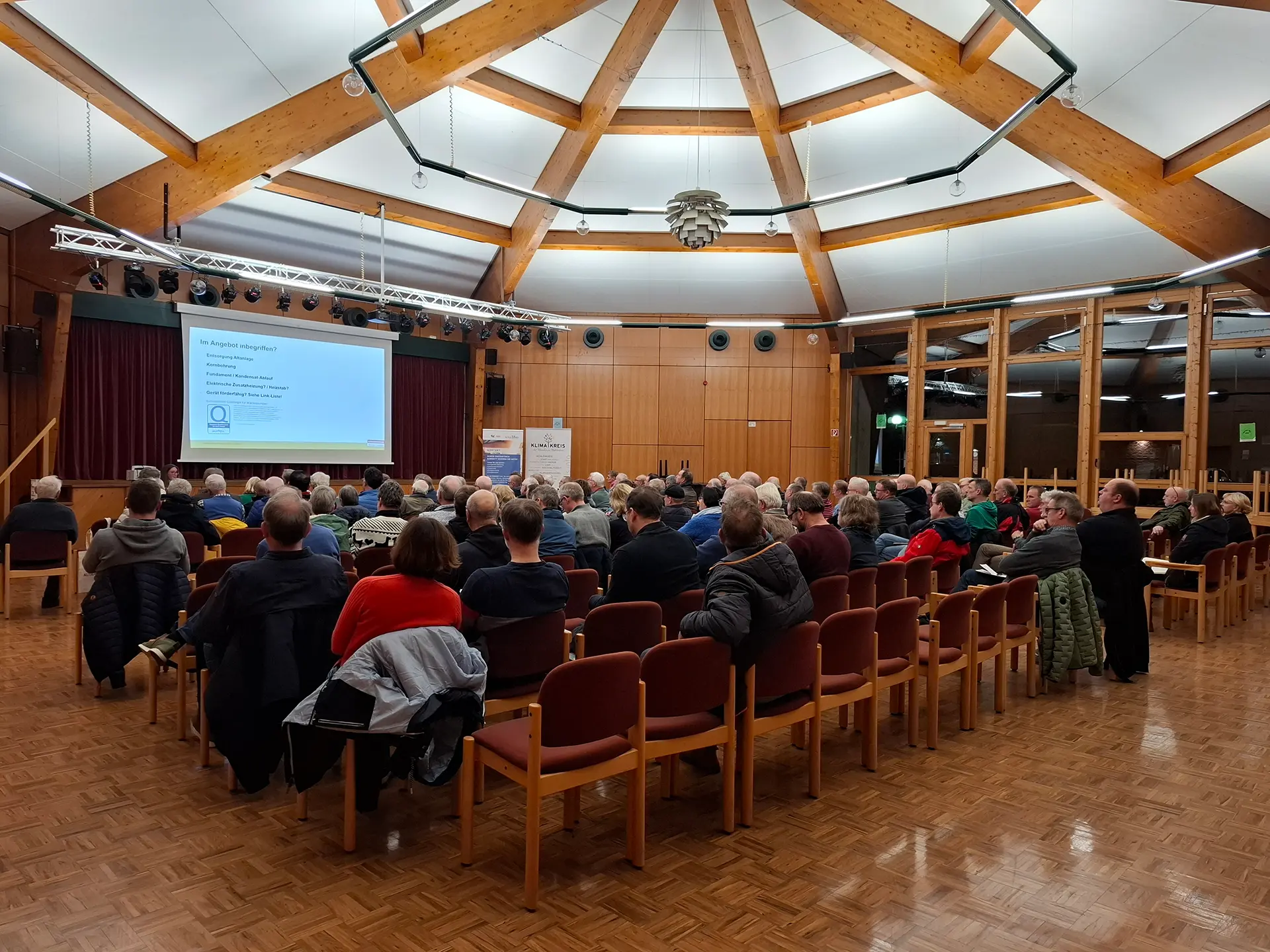 Gruppe von Menschen sitzt in einem Saal und blickt auf eine Leinwand mit Präsentationstext.